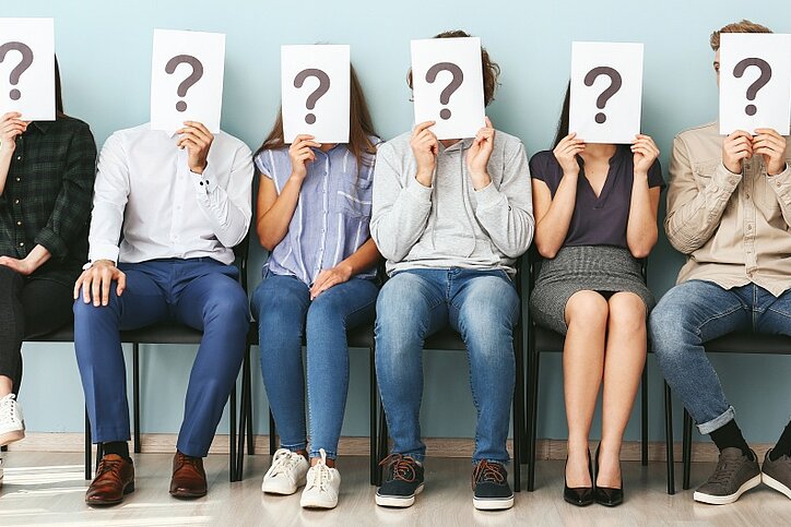 Junge Menschen verstecken ihre Gesichter hinter Papierblättern mit Fragezeichen, während sie in Innenräumen auf ein Vorstellungsgespräch warten.