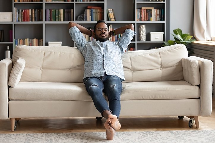 Mann sitzt mit hinter dem Kopf verschränkten Händen entspannt auf einem Sofa. © stock.adobe.com_fizkes