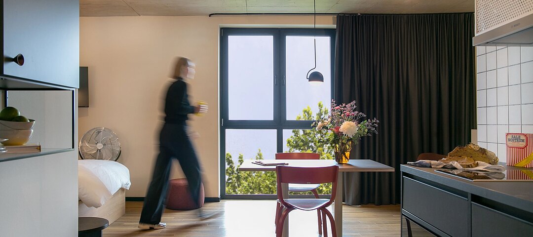 Stayery hat jetzt auch in der Ruhrstadt Bochum eine Adresse. Das Gebäude hatte Catella 2023 gekauft. © Stayery  Apartment mit Fenster, Tisch, Küchenzeile und einer Person, die durchläuft